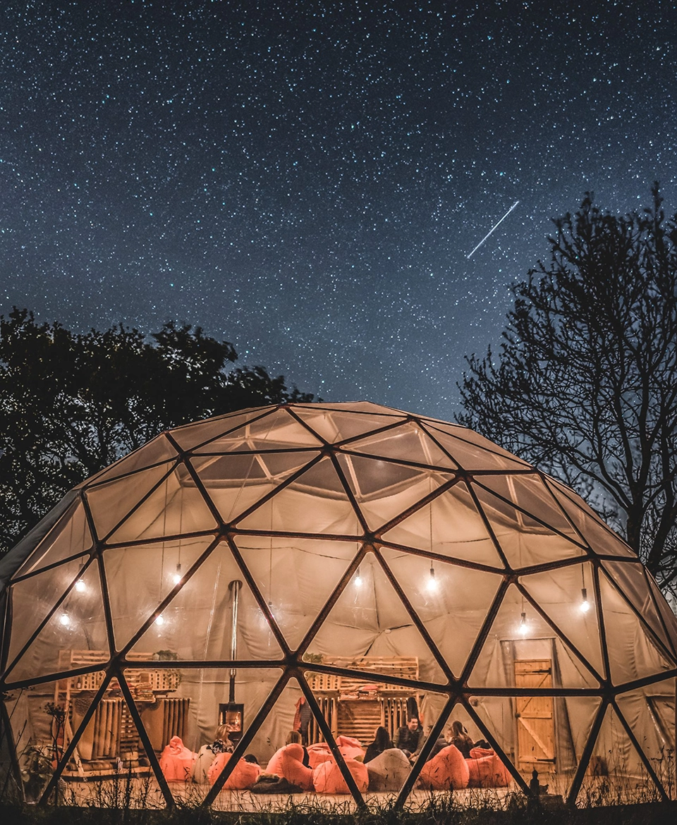 A large dome from TruDomes at night lit up with lights. Above is the night sky with lots of stars